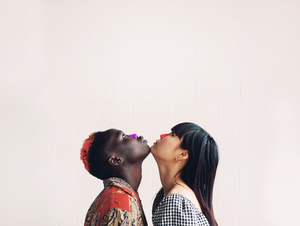 Two women facing each other and looking up. On the left the woman has black hair with a red stripe on it and purple sunscreen on her nose. On the right the woman also has black hair and orange sunscreen is on her nose.